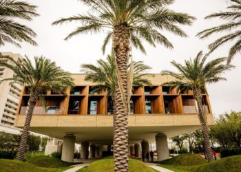 Modern building with large windows and a raised structure supported by columns, surrounded by palm trees and green grass beneath a cloudy sky. The International African American Museum Charleston stands out, with a tall apartment building in the background.