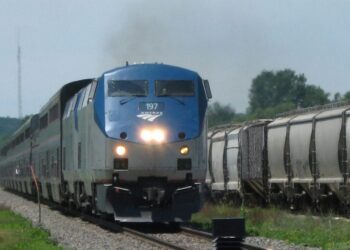 A blue and silver Amtrak passenger train travels on railroad tracks alongside several freight train cars on a clear, sunny day. Trees and greenery are visible in the background.
