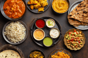 An assortment of Indian dishes including curry, yellow rice, naan, papadum, chutneys, dals, vegetable salad, and pakoras, all artistically arranged on a rustic brown table.