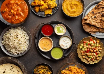 A variety of Indian foods on a dark table, including rice, curry, flatbread, grilled chicken, lentil stew, sauces, salad, and chutneys, all beautifully arranged in bowls and plates.