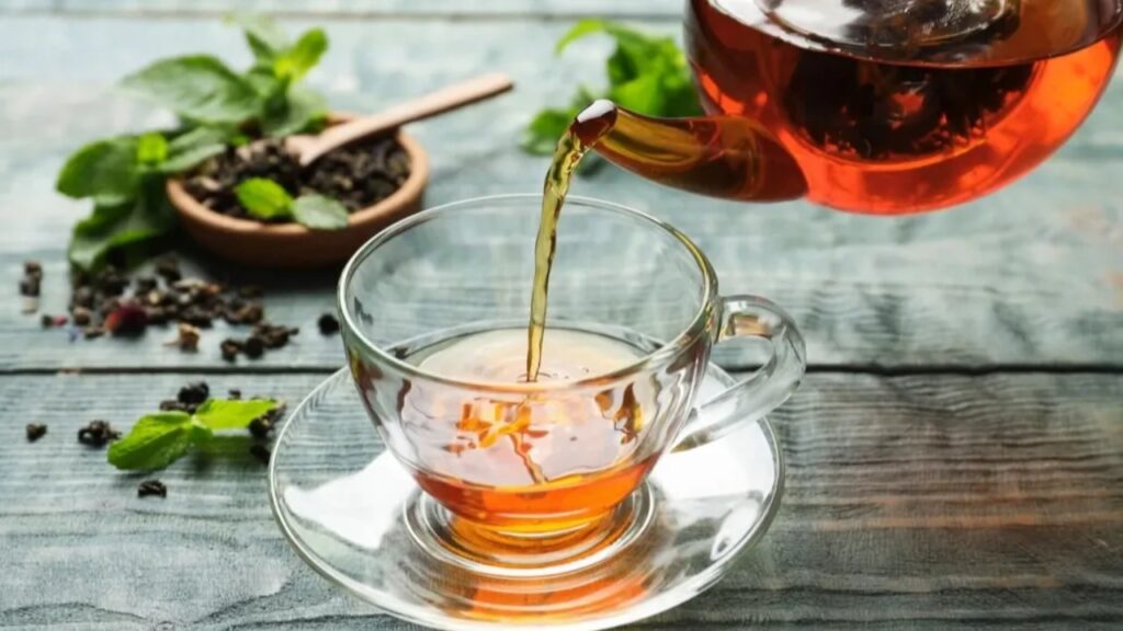 A glass teapot pours amber-colored tea into a clear teacup on a saucer. Loose tea leaves, a wooden spoon, and fresh mint leaves are arranged on a rustic wooden surface.
