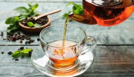 A glass teapot pours amber-colored tea into a clear teacup on a saucer. Loose tea leaves, a wooden spoon, and fresh mint leaves are arranged on a rustic wooden surface.