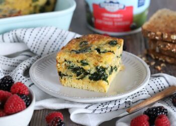 A slice of spinach and cheese egg casserole sits on a white plate, surrounded by a black-and-white striped napkin. Fresh berries and slices of whole grain bread are nearby on a wooden table.