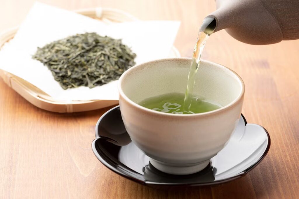 A cup of green tea, known for its health benefits, is being poured from a teapot into a ceramic cup on a black saucer, with loose green tea leaves on a tray in the background.