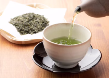 A cup of green tea, known for its health benefits, is being poured from a teapot into a ceramic cup on a black saucer, with loose green tea leaves on a tray in the background.
