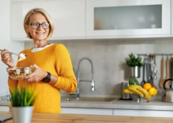A smiling woman wearing glasses and a yellow sweater stands in a modern kitchen, holding a bowl of salad and a fork—showcasing habits for long-term health. Fruit and kitchen utensils are visible in the background.