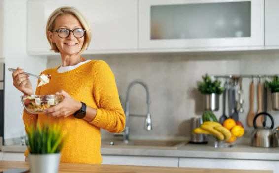 A smiling woman wearing glasses and a yellow sweater stands in a modern kitchen, holding a bowl of salad and a fork—showcasing habits for long-term health. Fruit and kitchen utensils are visible in the background.