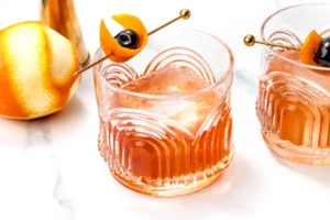 Two glasses of an alcohol-free old fashioned with large ice cubes, garnished with orange peel and a cherry on a gold skewer. An orange and a cocktail shaker are visible on a white marble surface.