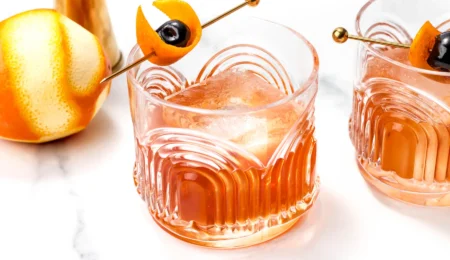 Two glasses of an alcohol-free old fashioned with large ice cubes, garnished with orange peel and a cherry on a gold skewer. An orange and a cocktail shaker are visible on a white marble surface.