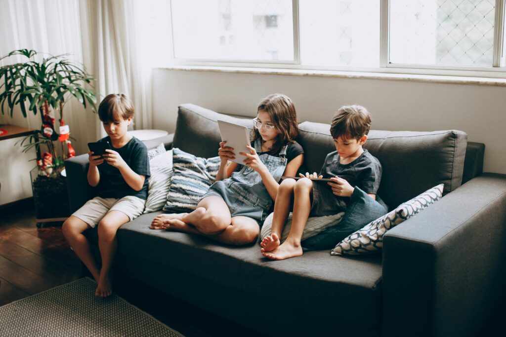 Three grandkids sit on a gray sofa in the bright living room, each enjoying their screen time—one with a smartphone, another with a tablet, and the third with a handheld console. Sunlight streams through large windows beside a leafy plant.