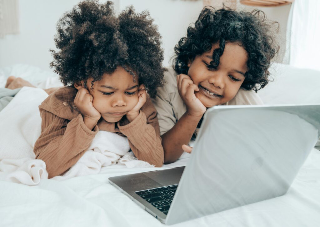 Two grandkids with curly hair lie on a bed, wrapped in blankets, looking intently at an open laptop during their screen time. One looks serious while the other smiles slightly.