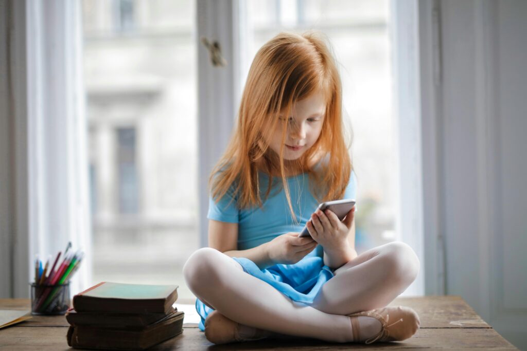 A young girl with long red hair, wearing a light blue dress and pink leggings, sits cross-legged on a table by a window, looking at a smartphone—a familiar scene for many grandkids and their daily screen time. Books and pencils sit beside her.