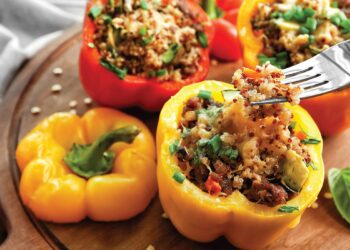 A close-up of colorful stuffed bell peppers filled with quinoa, vegetables, and herbs on a wooden board. A fork holds a bite of stuffing above a yellow pepper, with other peppers and garnish visible in the background.