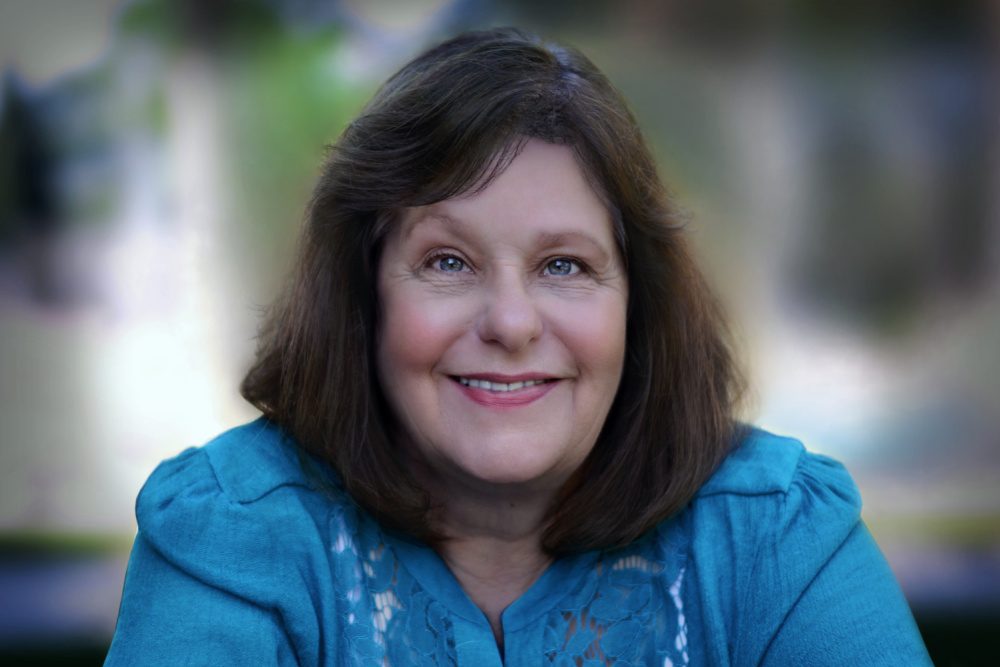 A middle-aged woman with shoulder-length brown hair and a blue blouse smiles at the camera. The background is softly blurred with green and neutral tones.
