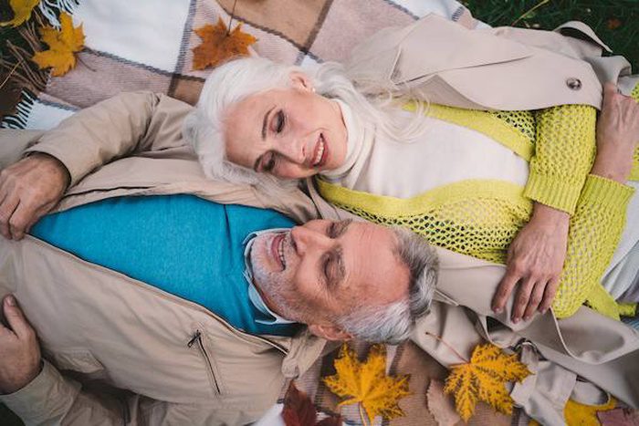 An older couple lies on a blanket outdoors, surrounded by autumn leaves. They are smiling and looking at each other, dressed warmly in coats and sweaters, enjoying a peaceful, affectionate moment together.