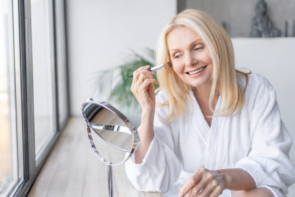 A smiling woman in a white robe sits by a window, carefully avoiding makeup that ages you as she applies product with a brush while looking into a round mirror on a wooden table.