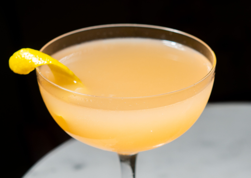 A coupe glass filled with a pale orange cocktail, garnished with a twist of lemon peel on the rim, set against a dark background.