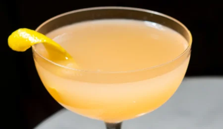 A coupe glass filled with a pale orange cocktail, garnished with a twist of lemon peel on the rim, set against a dark background.