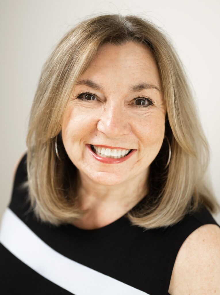 Lisa Copeland- image of A smiling woman with shoulder-length light brown hair, large hoop earrings, and a black sleeveless top with a white stripe poses in front of a neutral background, radiating the spirit of dating with confidence later in life.