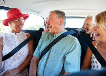 Four smiling seniors sit close together in the backseat of a car, wearing seatbelts. They are laughing and enjoying each others company, with sunlight coming through the windows.