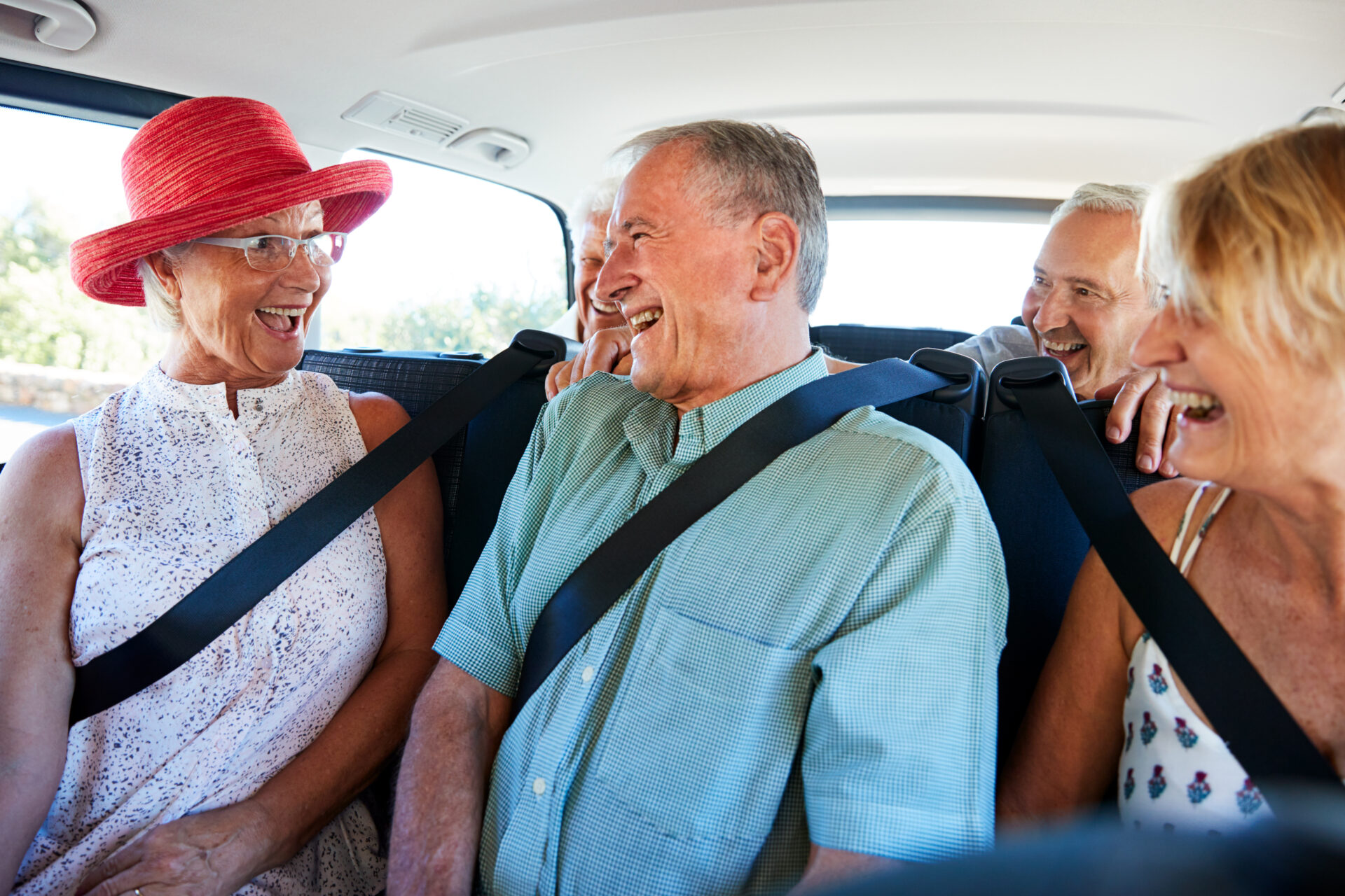 Four smiling seniors sit close together in the backseat of a car, wearing seatbelts. They are laughing and enjoying each others company, with sunlight coming through the windows.