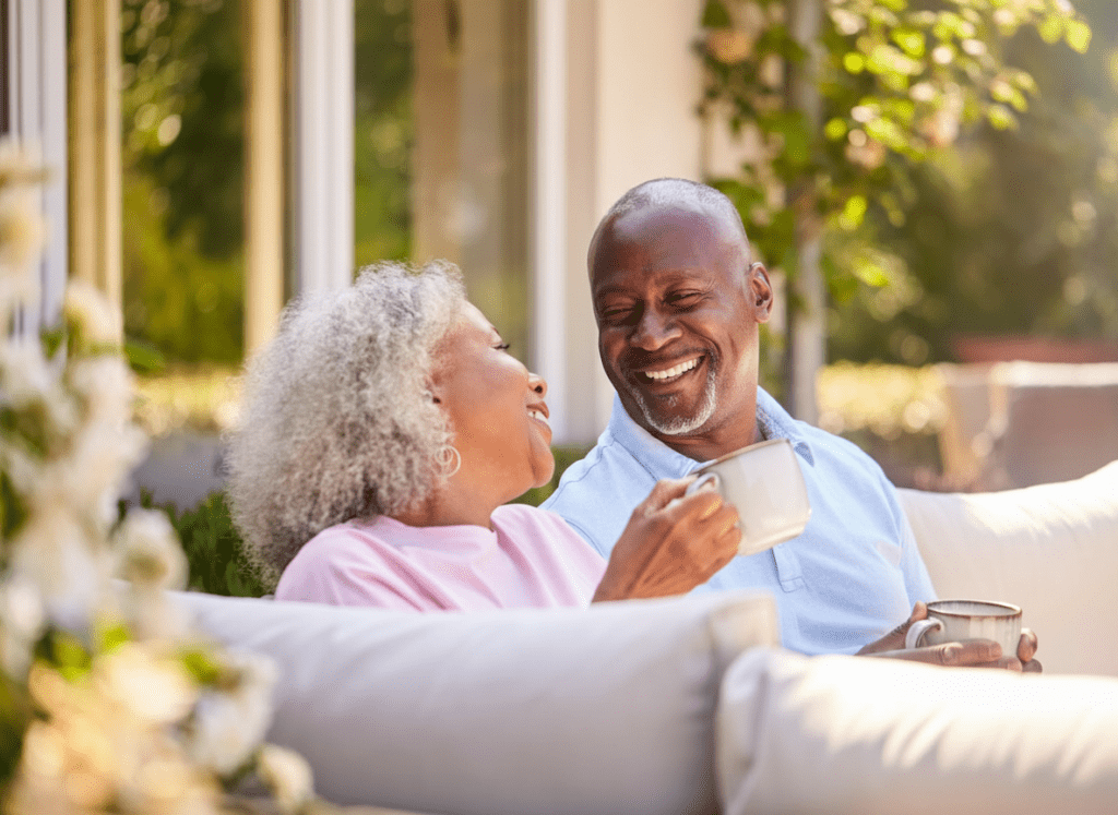 An older couple sits on an outdoor couch, smiling and enjoying hot drinks together in a sunlit garden. They appear happy and relaxed, surrounded by greenery and flowers.