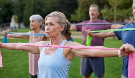 A group of older adults exercise outdoors, holding colorful resistance bands with arms extended. The focus is on a woman in front, with trees and blurred participants in the background.