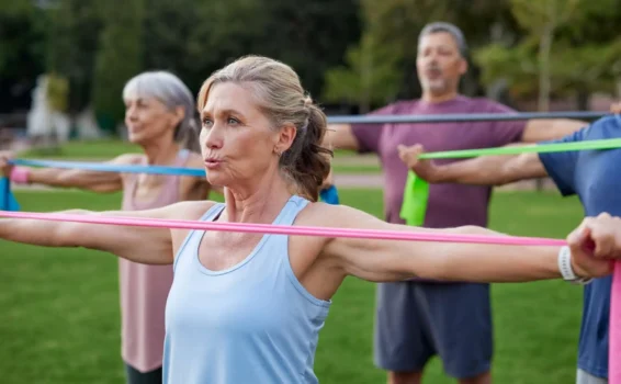 A group of older adults exercise outdoors, holding colorful resistance bands with arms extended. The focus is on a woman in front, with trees and blurred participants in the background.