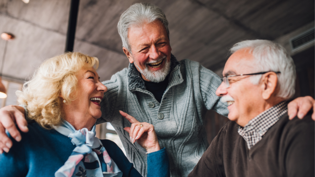 Three older adults are smiling and laughing together indoors. One man stands with his arms around a woman and another man, all appearing happy and enjoying each others company.
