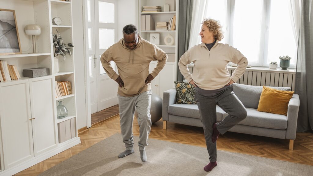 Two adults practicing balance exercises together in a cozy living room, each standing on one leg with hands on hips, smiling at each other. The room is bright with shelves, a couch, and a large window.