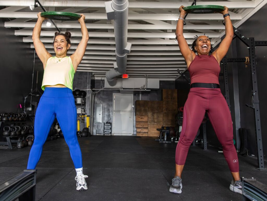 Two women in athletic wear smile and lift green weights overhead in a gym, standing side by side on a black floor with gym equipment and weights visible in the background.