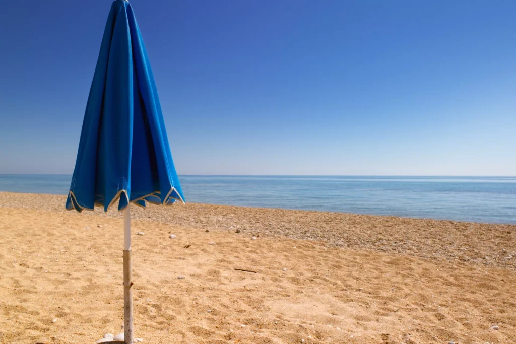 A closed blue beach umbrella stands on golden sand near a calm sea under a clear blue sky, with no people present.