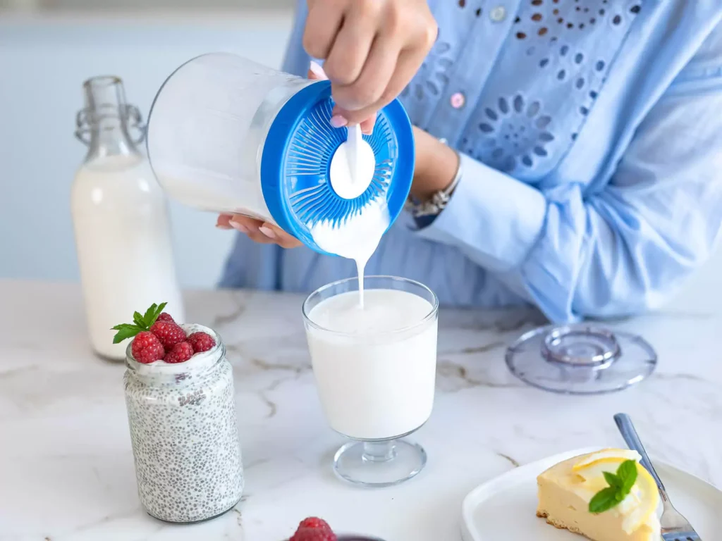 A person in a blue shirt pours yogurt from a container into a glass on a white table with a jar of chia pudding topped with raspberries and a plate with a dessert nearby.