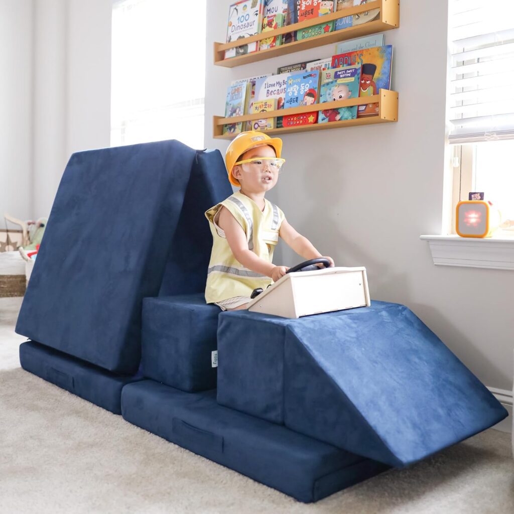 A young child wearing a construction vest and hard hat sits on blue foam cushions arranged like a vehicle, holding a toy steering wheel—surrounded by bookshelves and sunlight, this playful scene showcases creative kids room decor.
