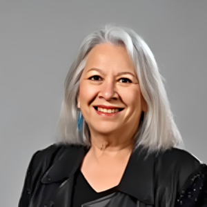 A smiling older woman with straight, shoulder-length gray hair, dressed in a black top and jacket, stands against a plain gray background—radiating confidence as she explores travel tips for seniors and new travel ideas and destinations.