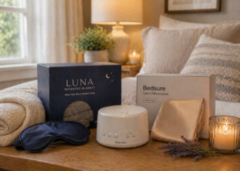 A cozy bedroom scene featuring a Luna weighted blanket box, a Bedsure satin pillowcase box with gold pillowcases, a black sleep mask, a white noise machine, a lit candle, a rolled blanket, and a sprig of lavender on a bed.