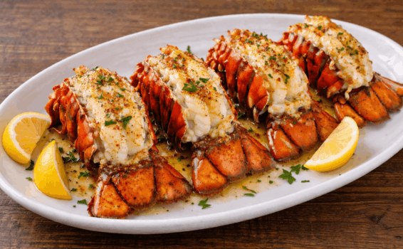 Four broiled lobster tails on a white oval plate, garnished with chopped parsley and accompanied by lemon wedges, with melted butter sauce drizzled on top. The plate rests on a wooden table.