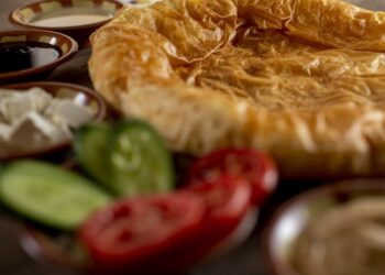 A close-up of a flaky, golden-brown pastry surrounded by fresh cucumber and tomato slices, white cheese cubes, and small bowls of assorted dips or sauces.