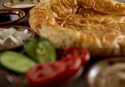 A close-up of a flaky, golden-brown pastry surrounded by fresh cucumber and tomato slices, white cheese cubes, and small bowls of assorted dips or sauces.