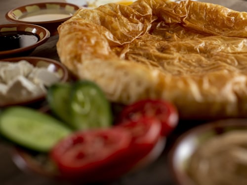 A close-up of a flaky, golden-brown pastry surrounded by fresh cucumber and tomato slices, white cheese cubes, and small bowls of assorted dips or sauces.