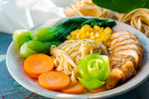 A bowl of noodles topped with sliced grilled chicken, bok choy, corn, carrot rounds, and a rolled cucumber garnish, arranged artfully on a light plate.
