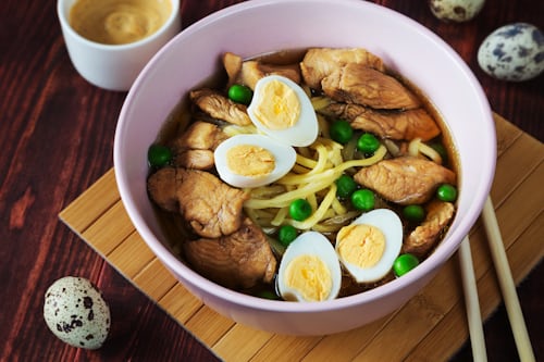 A bowl of noodle soup with pieces of chicken, halved boiled quail eggs, and green peas, served on a bamboo mat with chopsticks. A small cup of sauce and quail eggs are beside the bowl.