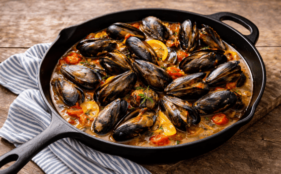 A cast-iron skillet filled with cooked mussels in a broth with cherry tomatoes, lemon slices, and herbs, resting on a blue and white striped cloth on a wooden surface.