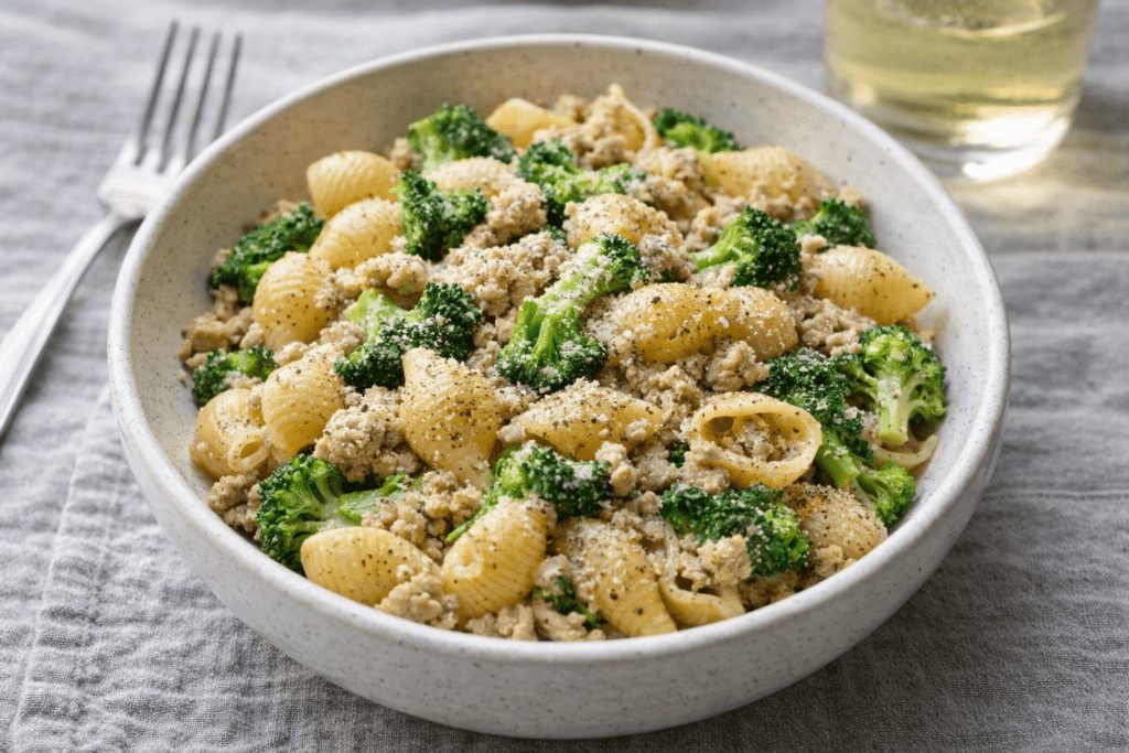 A bowl of pasta shells mixed with ground meat, broccoli florets, and sprinkled with black pepper, served on a gray cloth with a fork and a glass of white wine in the background.