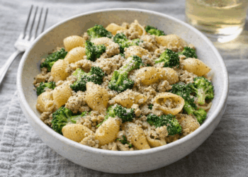 A bowl of pasta shells mixed with ground meat, broccoli florets, and sprinkled with black pepper, served on a gray cloth with a fork and a glass of white wine in the background.