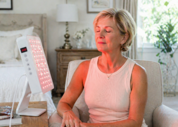 A woman sits in a cozy, well-lit room with her eyes closed, facing a tabletop red light therapy panel emitting red light onto her face. She appears relaxed and at ease.