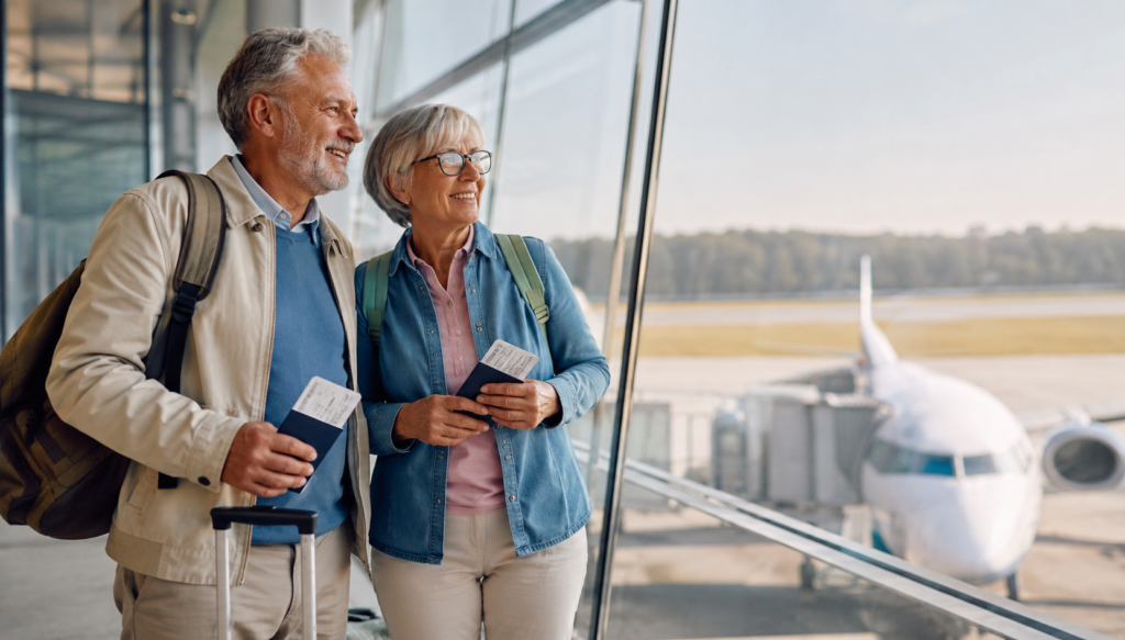 An older man and woman with backpacks stand by airport windows, holding passports and boarding passes, smiling as they look outside at a plane parked at the gate on a sunny day.