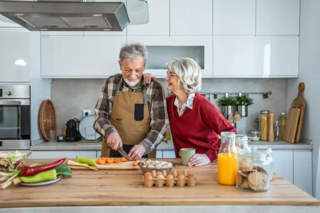 An elderly couple smiles together in a modern kitchen; embracing food as medicine, the man chops vegetables in an apron while the woman stands beside him with a mug. Eggs, juice, and baking ingredients are on the counter.