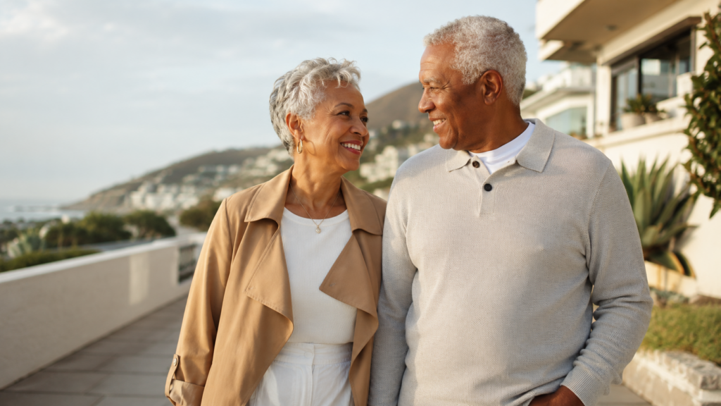 An older couple walks outdoors, smiling at each other. They are casually dressed and appear happy, with buildings, greenery, and hills in the background under a partly cloudy sky.