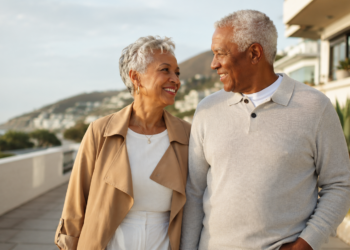 An older couple walks outdoors, smiling at each other. They are casually dressed and appear happy, with buildings, greenery, and hills in the background under a partly cloudy sky.